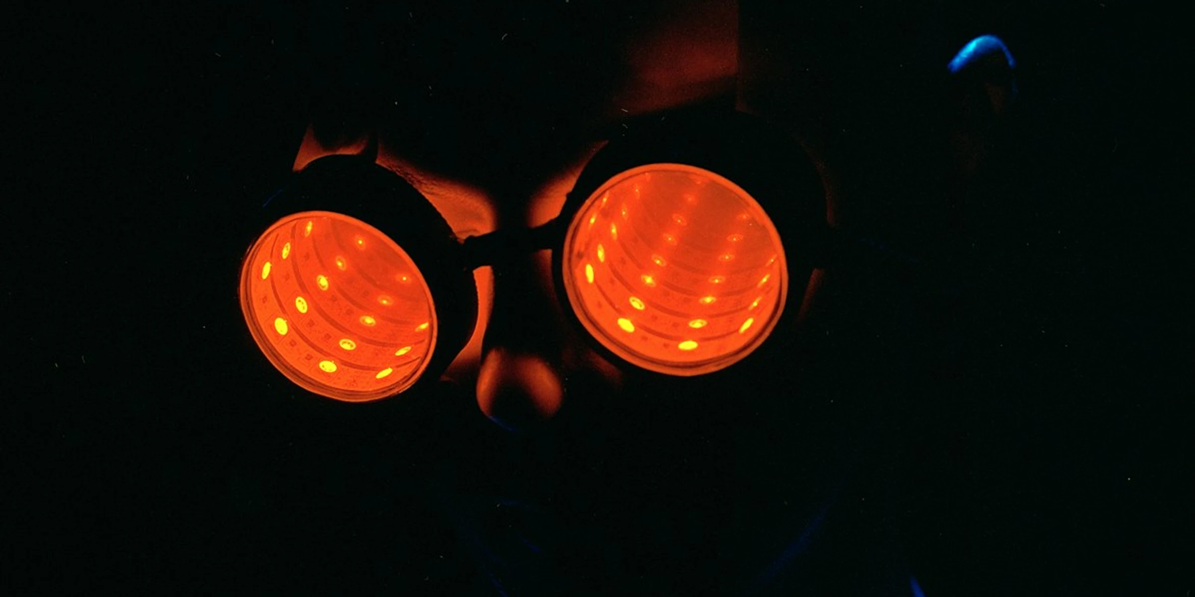 Close-up of a face in darkness wearing goggles whose two round lenses glow orange, lit from inside by concentric rings of small LEDs.